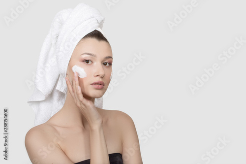 Portrait of a young woman with cream on her face. hand touch to the cheek. A towel on her head. White background