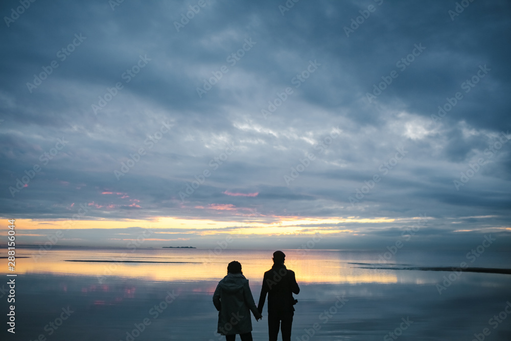 Silhouettes of couple man and woman holding hands near the sea or lake after sunset,  reflection of cloudy sky in water surface.