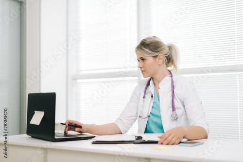 Young woman doctor woman working on laptop in office. Girl doctor makes a note in the outpatient card