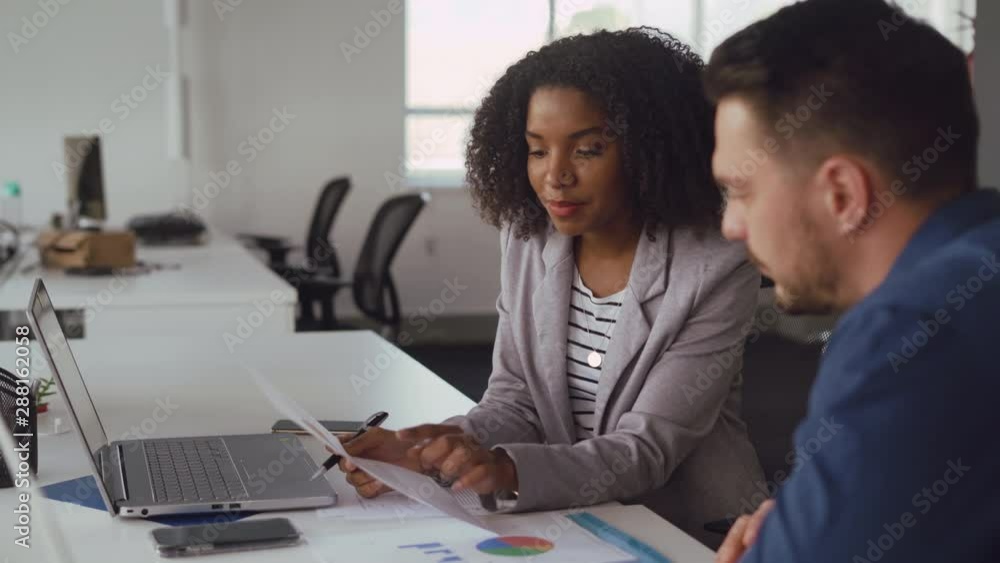 Young smiling african businesswoman explaining new ideas and plans to male colleague holding chart in hand at desk in office Young smiling african businesswoman explaining new ideas and plans to male colleague holding chart in hand at desk in office