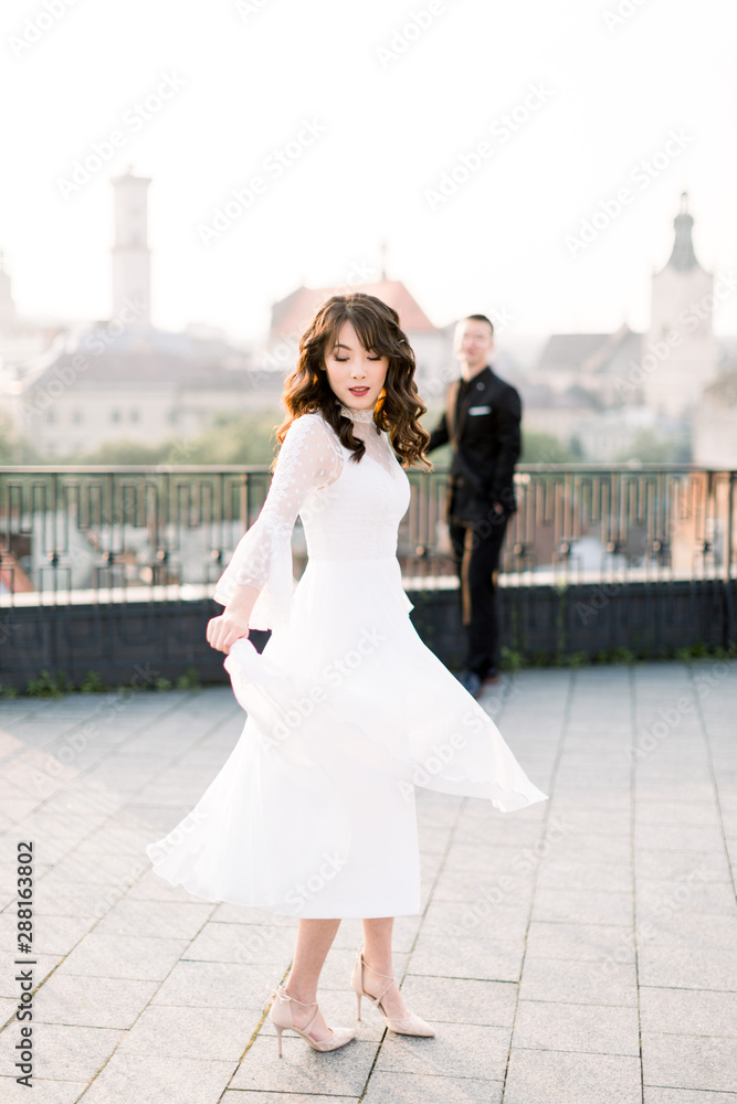 Obraz premium Young beautiful Asian bride in white wedding dress dancing on the terrace of ancient city. Groom standing on the background. Wedding walk in old city