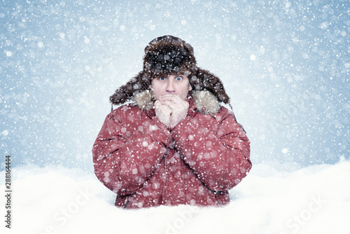 Frozen man in red winter clothes stands waist-deep in a snowdrift and warms his hands, cold, snow, frost, blizzard 
