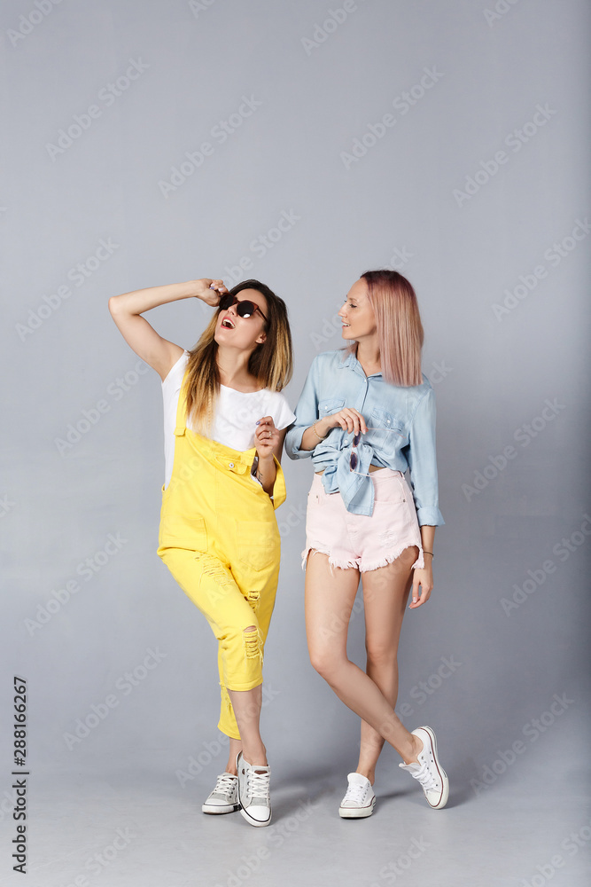Shot of cheerful young female best friends in sunglasses with pleasant appearance and broad smiles in stilysh color jeans casual clothes having fun over gray background. Two overjoyed student girls Shot of cheerful young female best friends in sunglasses with pleasant appearance and broad smiles in stilysh color jeans casual clothes having fun over gray background. Two overjoyed student girls
