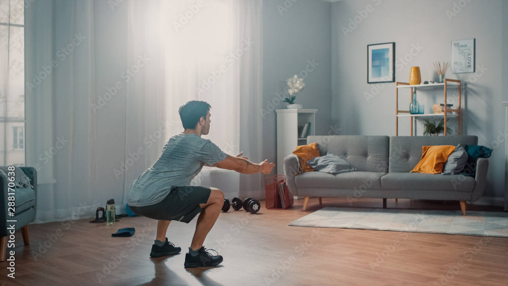 Strong Athletic Fit Man in T-shirt and Shorts is Doing Squat Exercises at Home in His Spacious and Bright Apartment with Minimalistic Interior. Strong Athletic Fit Man in T-shirt and Shorts is Doing Squat Exercises at Home in His Spacious and Bright Apartment with Minimalistic Interior.