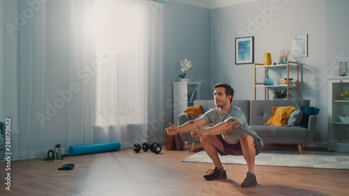 Muscular Athletic Fit Man in T-shirt and Shorts is Doing Squat Exercises at Home in His Spacious and Bright Apartment with Minimalistic Interior.