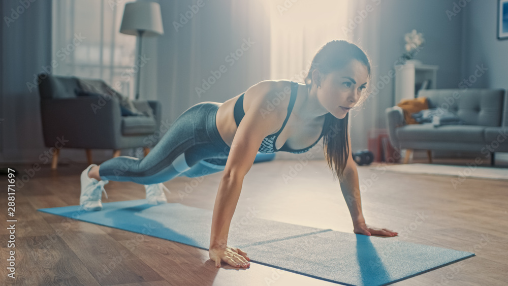 Strong Confident Beautiful Fitness Girl in Grey Athletic Sportswear is Doing Push Up Workout Exercises in Her Bright and Spacious Apartment with Cozy Minimalistic Interior. Strong Confident Beautiful Fitness Girl in Grey Athletic Sportswear is Doing Push Up Workout Exercises in Her Bright and Spacious Apartment with Cozy Minimalistic Interior.