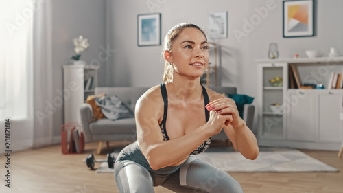Strong and Fit Beautiful Girl in an Athletic Top is Doing Squat Exercises in Her Bright and Spacious Living Room with Minimalistic Interior.