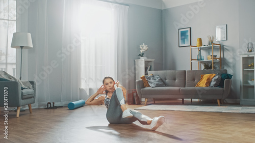 Strong and Fit Beautiful Girl in a Grey Athletic Outfit Energetically Exercising in Her Bright and Spacious Living Room with Minimalistic Interior. photo