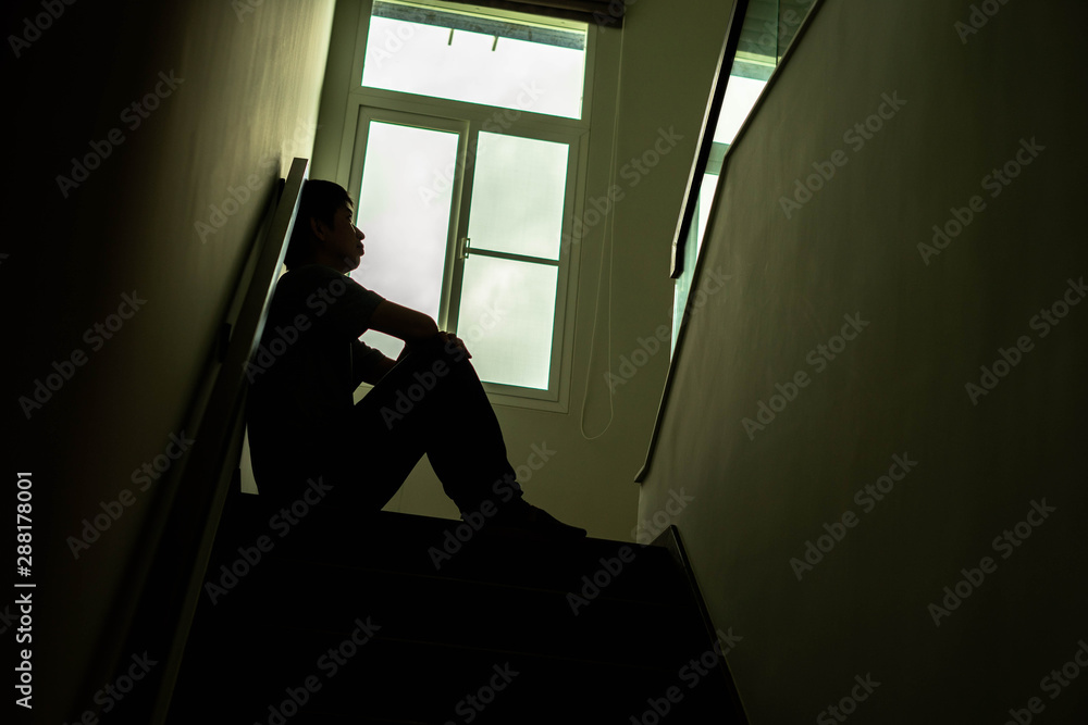 Silhouette of a sad young man Sitting at the stairs in the dark leaning against the wall with his back, The sun is in sorrow, Life problems, illness.