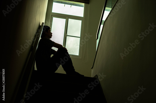 Silhouette of a sad young man Sitting at the stairs in the dark leaning against the wall with his back, The sun is in sorrow, Life problems, illness.