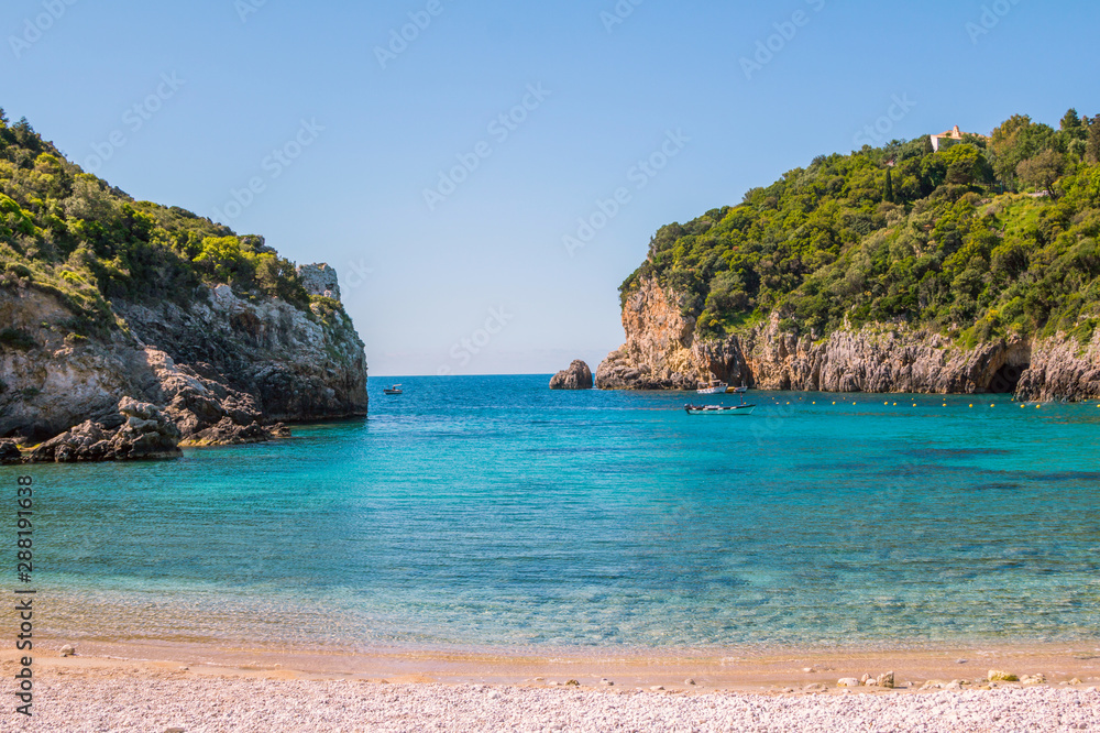 Seascape – lagoon with turquoise water, mountain with cliffs, green trees, blooming bushes, rocks in a blue water, colorful cruise touristic boats. Corfu Island, Greece. Seascape – lagoon with turquoise water, mountain with cliffs, green trees, blooming bushes, rocks in a blue water, colorful cruise touristic boats. Corfu Island, Greece.