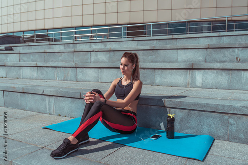 tanned woman, enjoying music headphones, listens podcast online app on Internet. Female athlete sits steps, resting jogging morning on run, summer city, girl doing fitness, youth lifestyle, healthy.