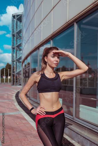 girl in summer in city, sportswear on run in morning closes her palm from bright sun, looks into distance. Search for friends girlfriends at a fitness workout , lifestyle healthy way of thinking.