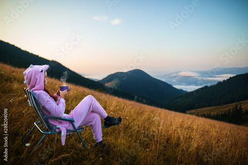 Young girl in a pink dragon costume drinking coffee while sitting in a chair on top of a mountain. Incredible mountain landscape at sunrise. Girl in pink pajamas drinks hot drink in the mountains