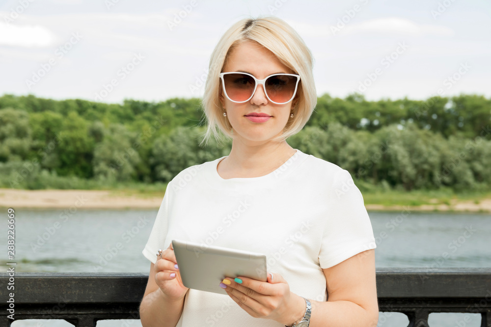 A young girl with glasses stands outdoors in a park near the river, holds a tablet.