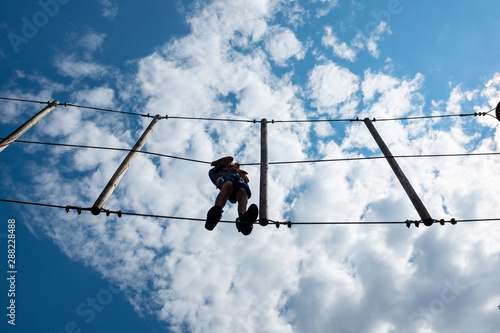 Boy climbing rope trail viewed from bottom in an adventure park. Silhouettes in front of a cloudy sky background.