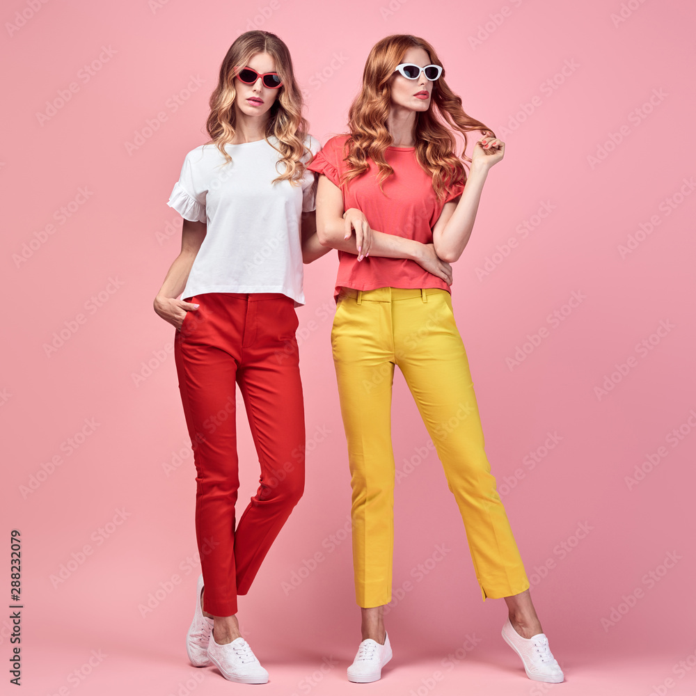 Fashion. Two Beautiful woman in Trendy summer outfit posing on coral. Graceful sisters friends in yellow coral pants with curly hair, make up. Colorful creative fashionable Indoor shot