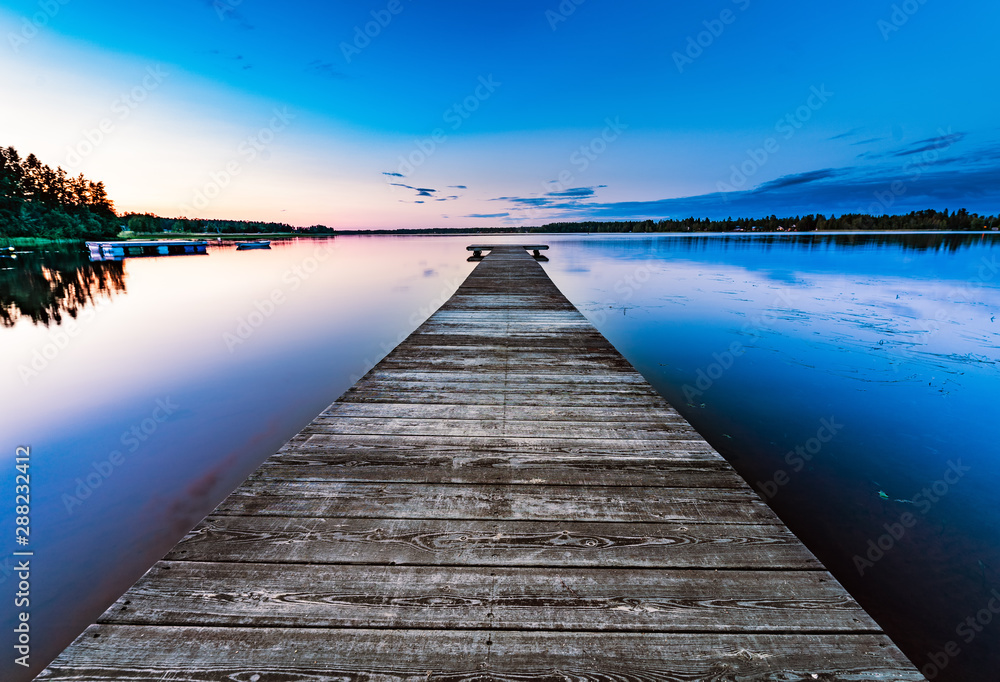 Very very long wooden bridge, almost to horizon, on the calm lake, summer sunset - blue rose skies reflected in still water. Northern Sweden