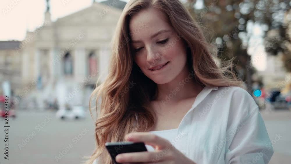 Smiling young woman with curly red hair walks down the street and uses mobile phone chats with friends feel happy Smiling young woman with curly red hair walks down the street and uses mobile phone chats with friends feel happy