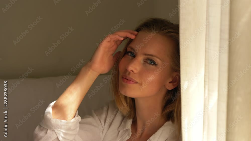 Blonde Female Sitting in White Bedroom Smiling by the Curtain Blonde Female Sitting in White Bedroom Smiling by the Curtain