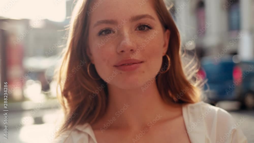 Close-up portrait of young charming woman with curty hair and freckles looking and smiling at camera at city center sun shines at the background Close-up portrait of young charming woman with curty hair and freckles looking and smiling at camera at city center sun shines at the background
