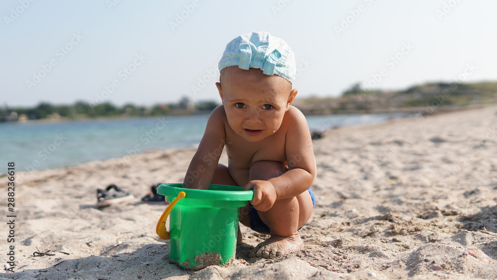 Little boy squatting on sea beach and taking sand in pail in his hand Little boy squatting on sea beach and taking sand in pail in his hand