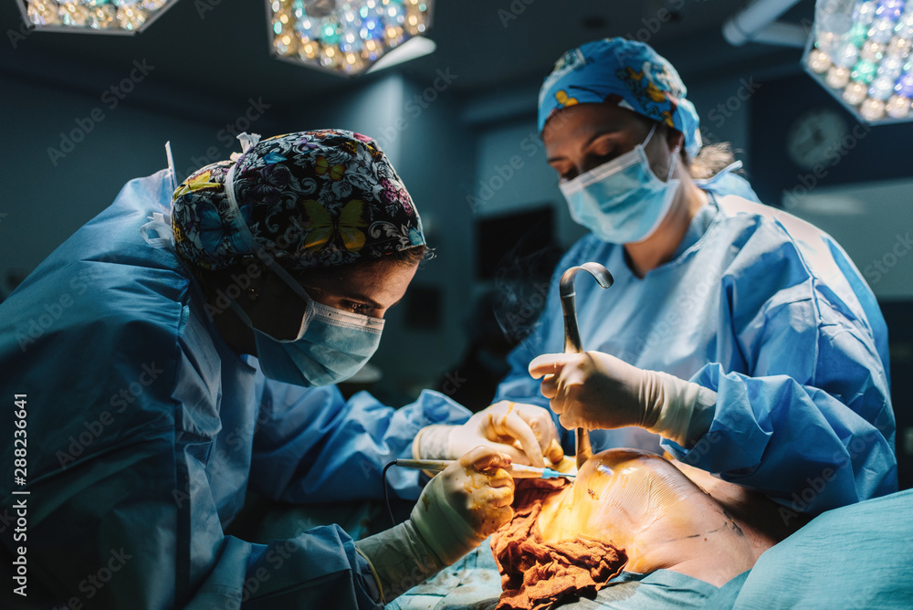 Side view of serious young doctor in protective mask and cap making surgery with instruments and crop nurse Side view of serious young doctor in protective mask and cap making surgery with instruments and crop nurse