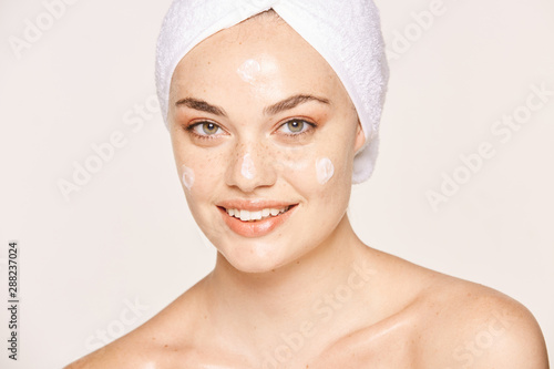Positive freckled woman with towel on head and healthy skin taking care of face with cream and looking at camera isolated on white background photo