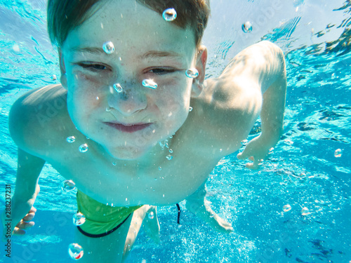 Squinting boy in shorts swimming under water of pool holding breath and looking at camera photo