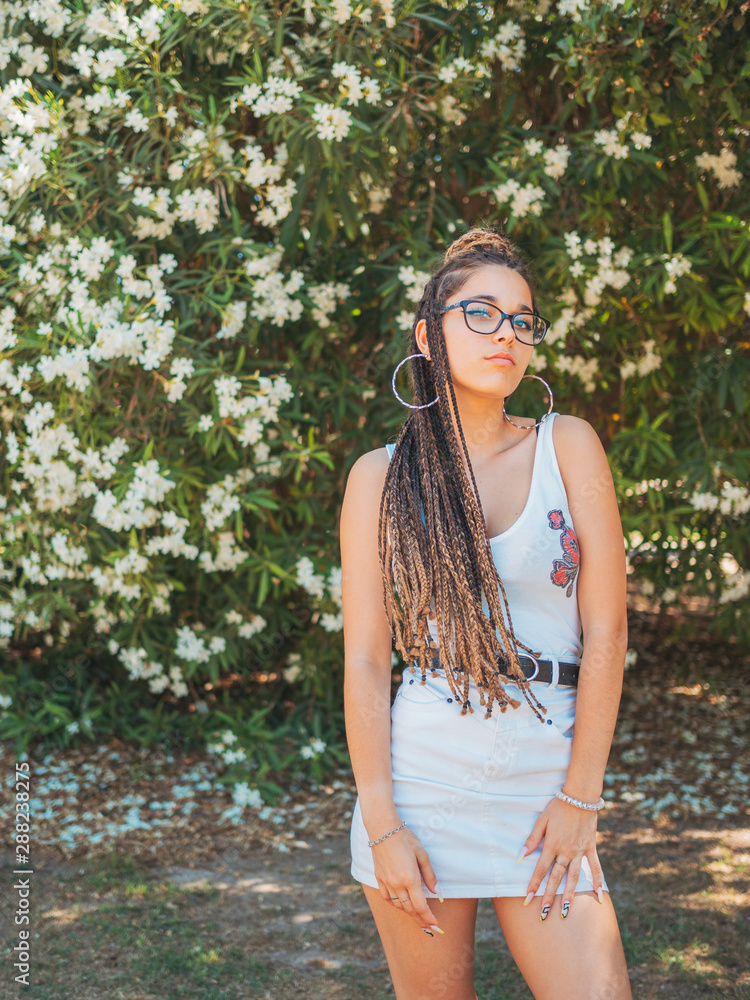 Beautiful young woman in glasses summer clothes and with dreadlocks standing near blooming trees and looking at camera Beautiful young woman in glasses summer clothes and with dreadlocks standing near blooming trees and looking at camera