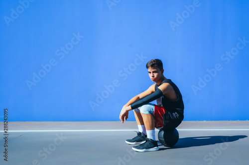 Side view of a man sitting on basket ball in front of a blue wall.