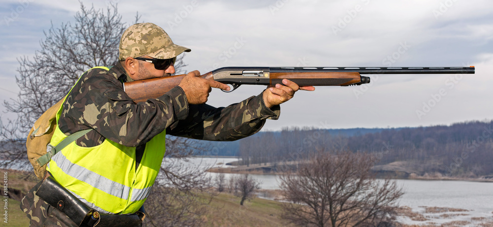 Fototapeta premium Hunting period, autumn season open. A hunter with a gun in his hands in hunting clothes in the autumn forest in search of a trophy. A man stands with weapons and hunting dogs tracking down the game. 