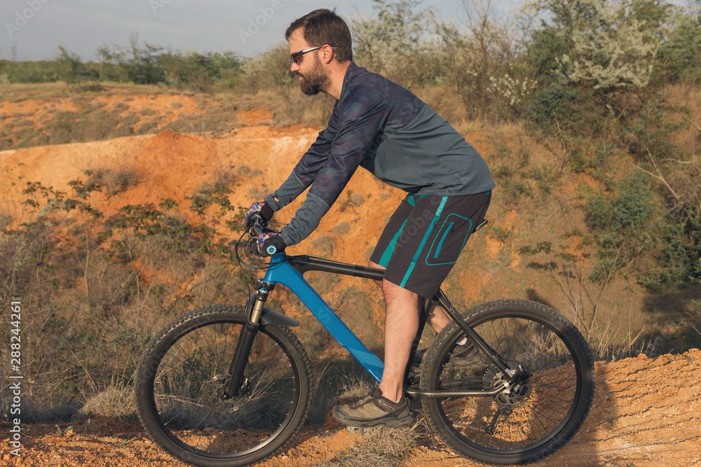 Cyclist in shorts and jersey on a modern carbon hardtail bike with an air suspension fork rides off-road on the orange-red hills at sunset evening in summer Cyclist in shorts and jersey on a modern carbon hardtail bike with an air suspension fork rides off-road on the orange-red hills at sunset evening in summer