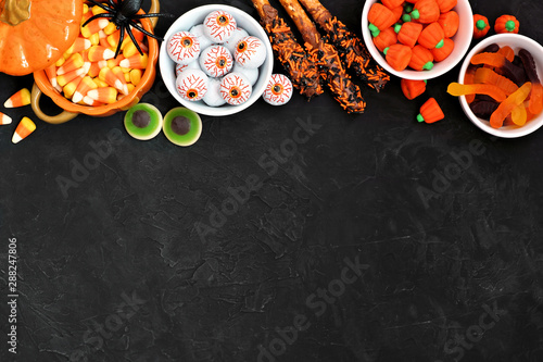 Halloween candy top border over a black stone background with copy space. Assortment of sweet, spooky treats. Above view. Buffet party food concept.