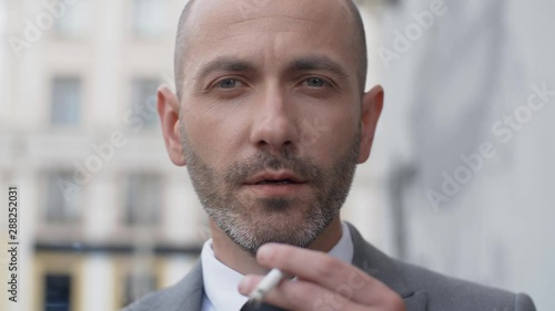 Close up attractive middle aged bearded man in business suit in a good mood smoke cigarette in front of camera in the street outdoors. Portrait of handsome male businessman standing on office porch photo