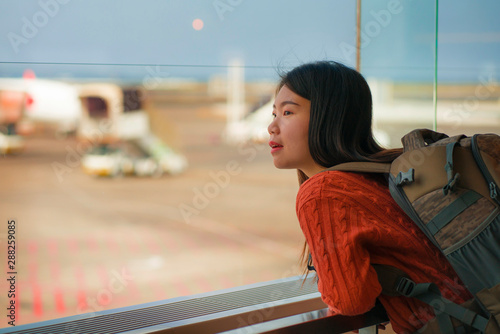 young happy and excited Asian Korean student woman with backpack at airport departure lounge watching aircraft through glass window smiling ready for boarding