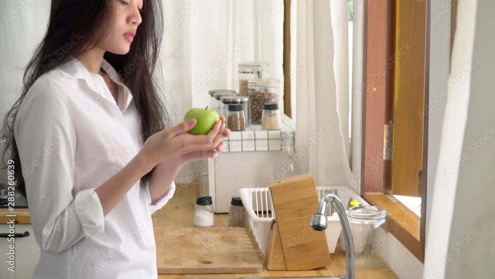 4K Medium Crane shot of lonely young asian woman standing in kitchen with holding green apple. Unhappy asian teenage girl standing alone near kitchen window thinking and looking away with sadness. 4K Medium Crane shot of lonely young asian woman standing in kitchen with holding green apple. Unhappy asian teenage girl standing alone near kitchen window thinking and looking away with sadness.