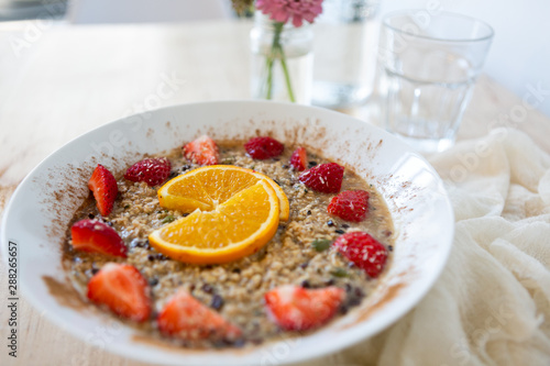 Superfood breakfast oatmeal bowl with fresh strawberries, orange slices, and maple syrup, flowers on table, wooden background