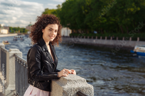 Young beautiful happy girl with curly hair is standing on the embankment and looking at the city. Woman with a departure in a leather jacket standing by the river