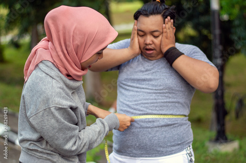 Two people, muslim woman and fat man, proportional and over weight, comparing themselves, by measure the circumference of the stomach with a meter