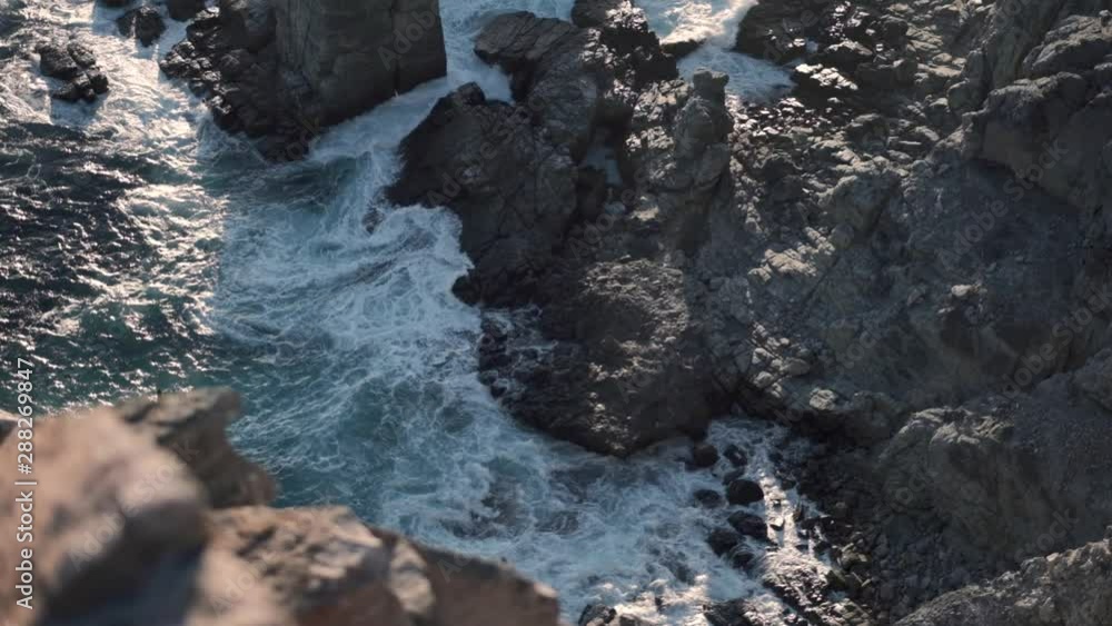 Tilt shot from above, of a rocky coastline with waves crashing on the shore, on a sunny evening Tilt shot from above, of a rocky coastline with waves crashing on the shore, on a sunny evening