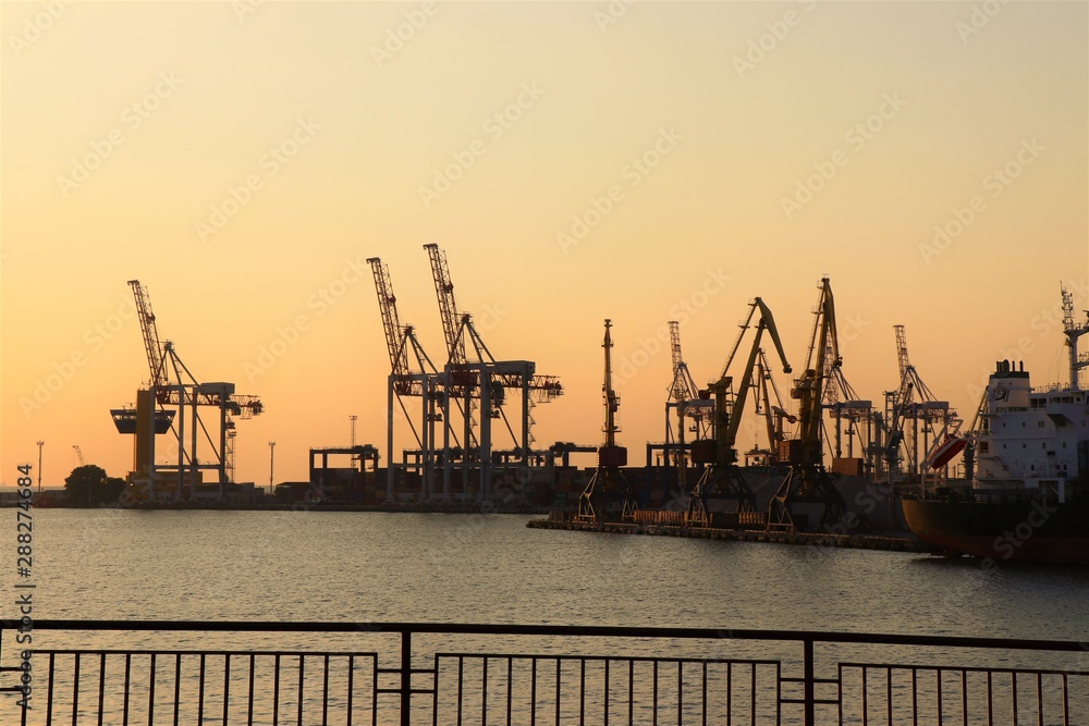 Cranes work with containers, in the commercial port of Odessa. In the biggest harbor of the Black sea. Photo taket during golden hours. Colorful sky and Black sea in the background.