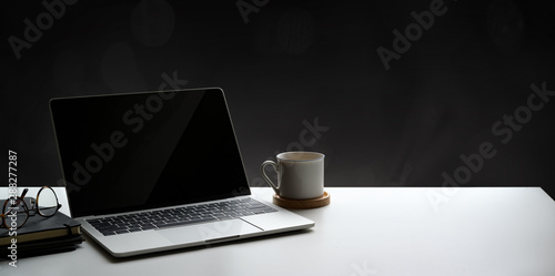 Open laptop computer with coffee cup and notebook on white table and blank wall background