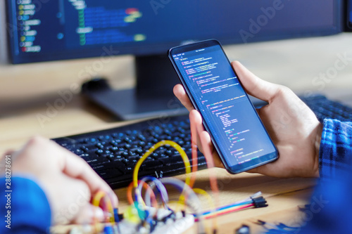 Developer is connecting breadboard to microcontroller. Man is holding smartphone with program code software for controlling electronic device. Chips, resistors, diodes on desktop of hardware engineer. photo