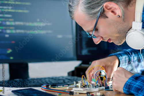 Developer is connecting breadboard to microcontroller for creating prototype of electronic device. Chips, resistors, light-emitting diodes on desktop of hardware engineer. Program code on background.
