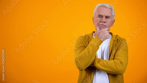 Senior man touching chin, thinking about decision against orange background photo
