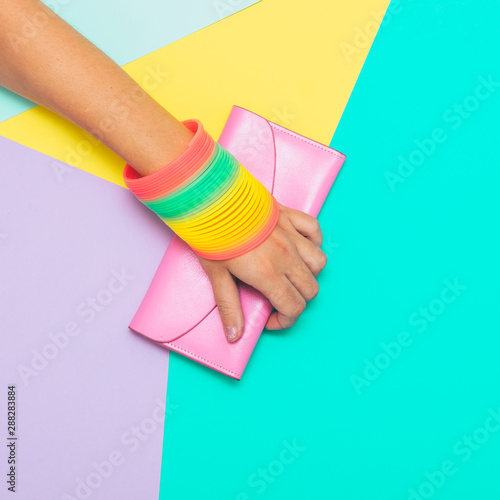 Hand with rainbow accessories holds pink purse on geometric background
