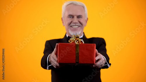 Cheerful male retiree in suit presenting red giftbox on camera, birthday present © motortion