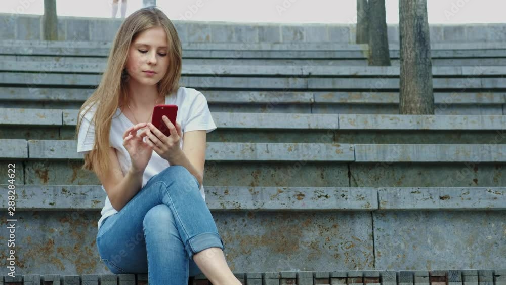 Young beautiful girl in a park holds a smartphone in her hands, writes a message. Leafing through photos on the phone, chatting in social networks with friends. Sits on concrete. Young beautiful girl in a park holds a smartphone in her hands, writes a message. Leafing through photos on the phone, chatting in social networks with friends. Sits on concrete.