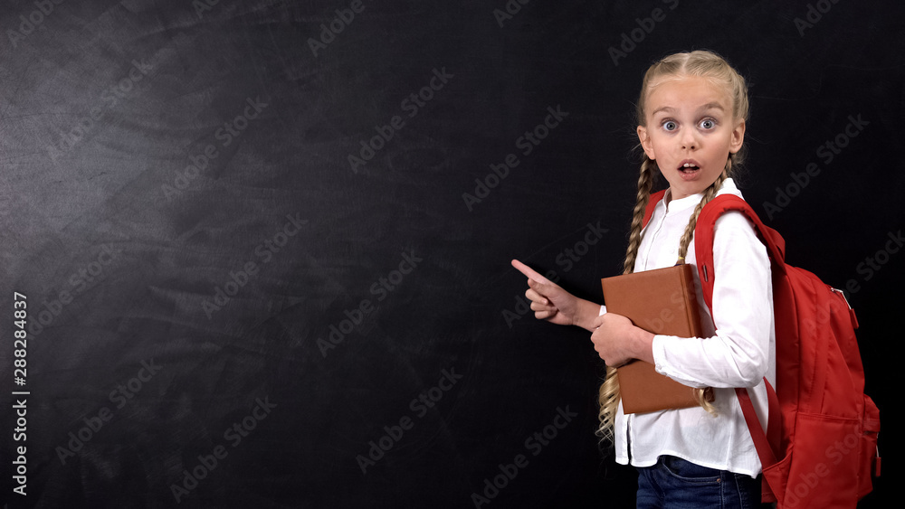 Shocked female kid with rucksack holding book, pointing finger at blackboard Shocked female kid with rucksack holding book, pointing finger at blackboard